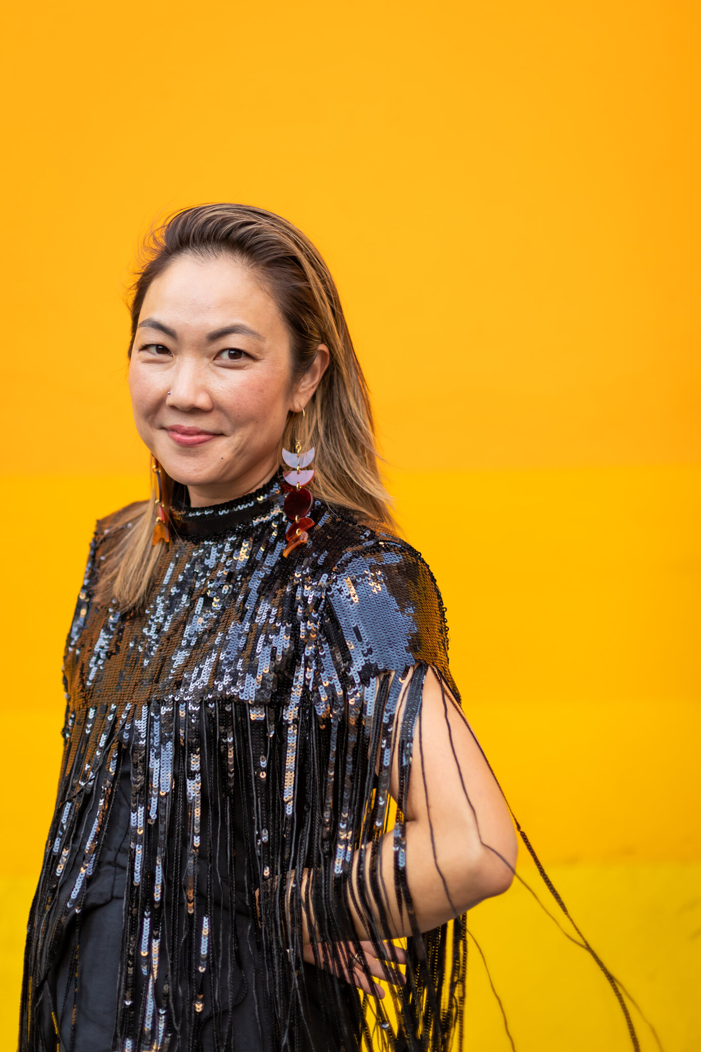 A person stands in front of a bright yellow-orange wall, wearing a black sequined top with long fringe and large earrings. They have long hair and are smiling slightly.
