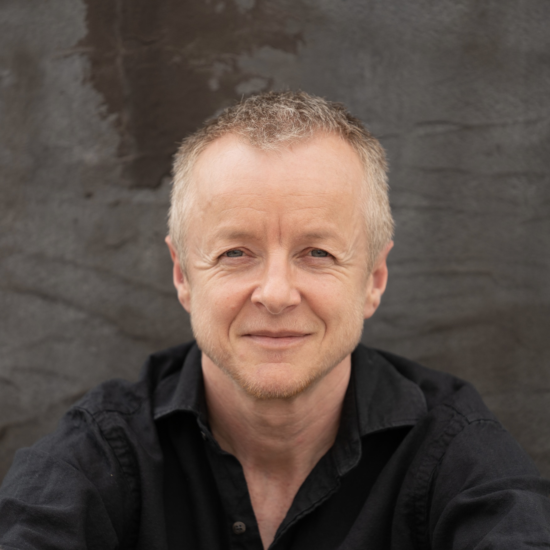 A person with short, light-colored hair and a slight smile is wearing a dark shirt and posing against a textured gray background.