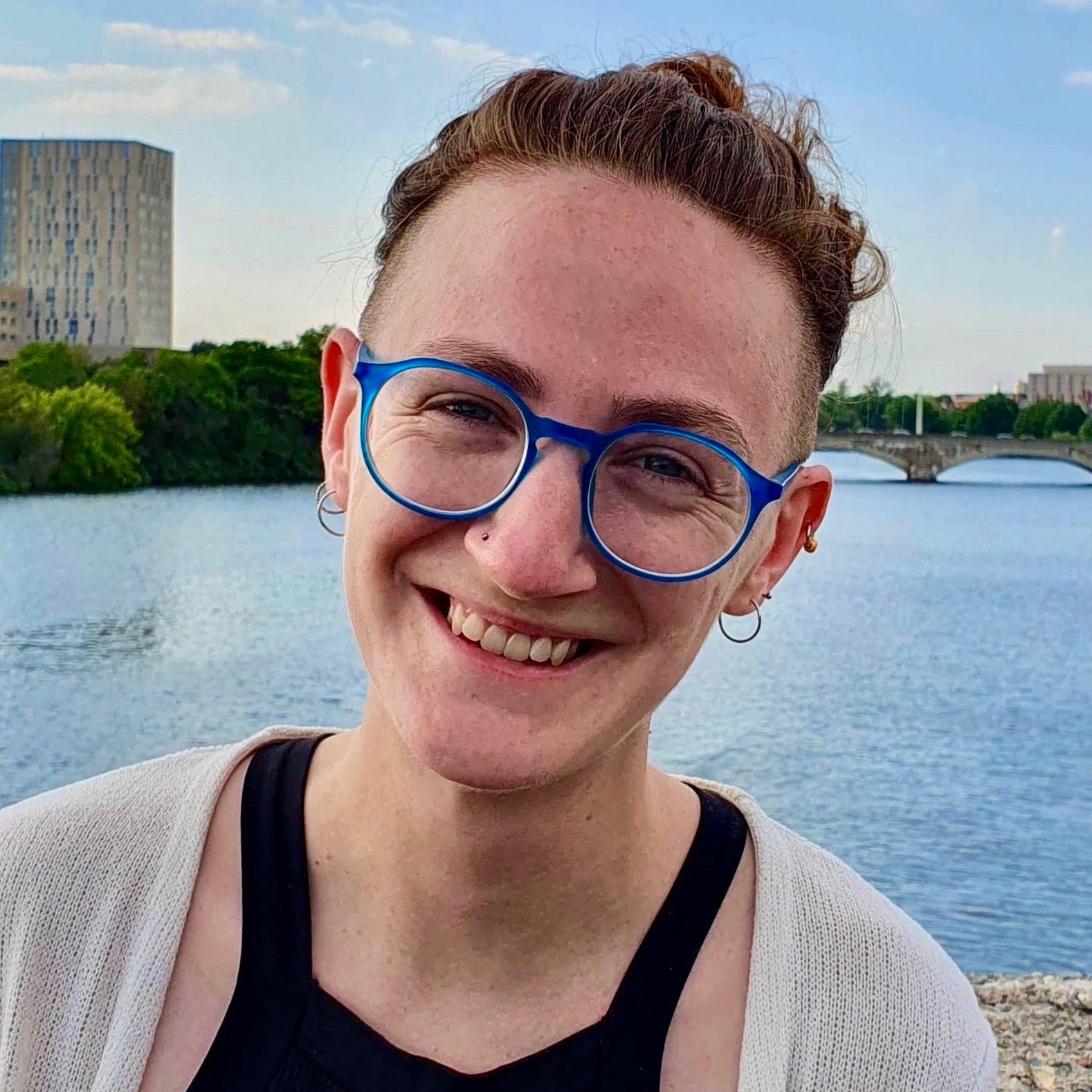 A person with short hair and blue-rimmed glasses smiles by a river. They are wearing a black top and a light cardigan. A bridge and tree-lined riverbank are visible in the background.