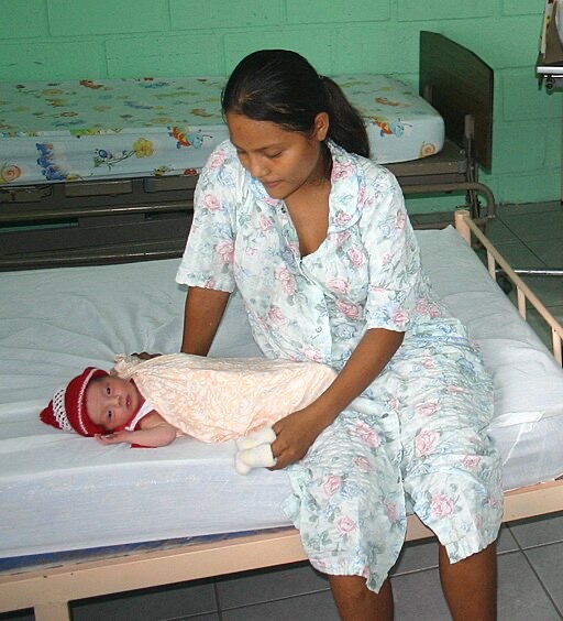 A woman in a floral dress sits on a hospital bed, gently tending to a baby wrapped in a blanket with a red hat. Another bed with a patterned sheet is in the background against a green wall.