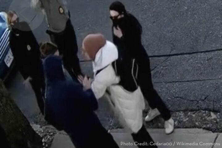 A group of masked individuals confront each other on a street. One person, wearing a long white coat and brown cap, gestures towards a person in a dark hoodie. Two others stand nearby, observing. The scene appears tense.