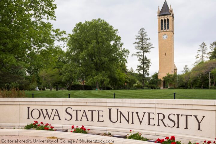 Campus scene featuring the Marston Water Tower and lush green trees at Iowa State University with Iowa State University written on a stone sign adorned with red flowers in the foreground.