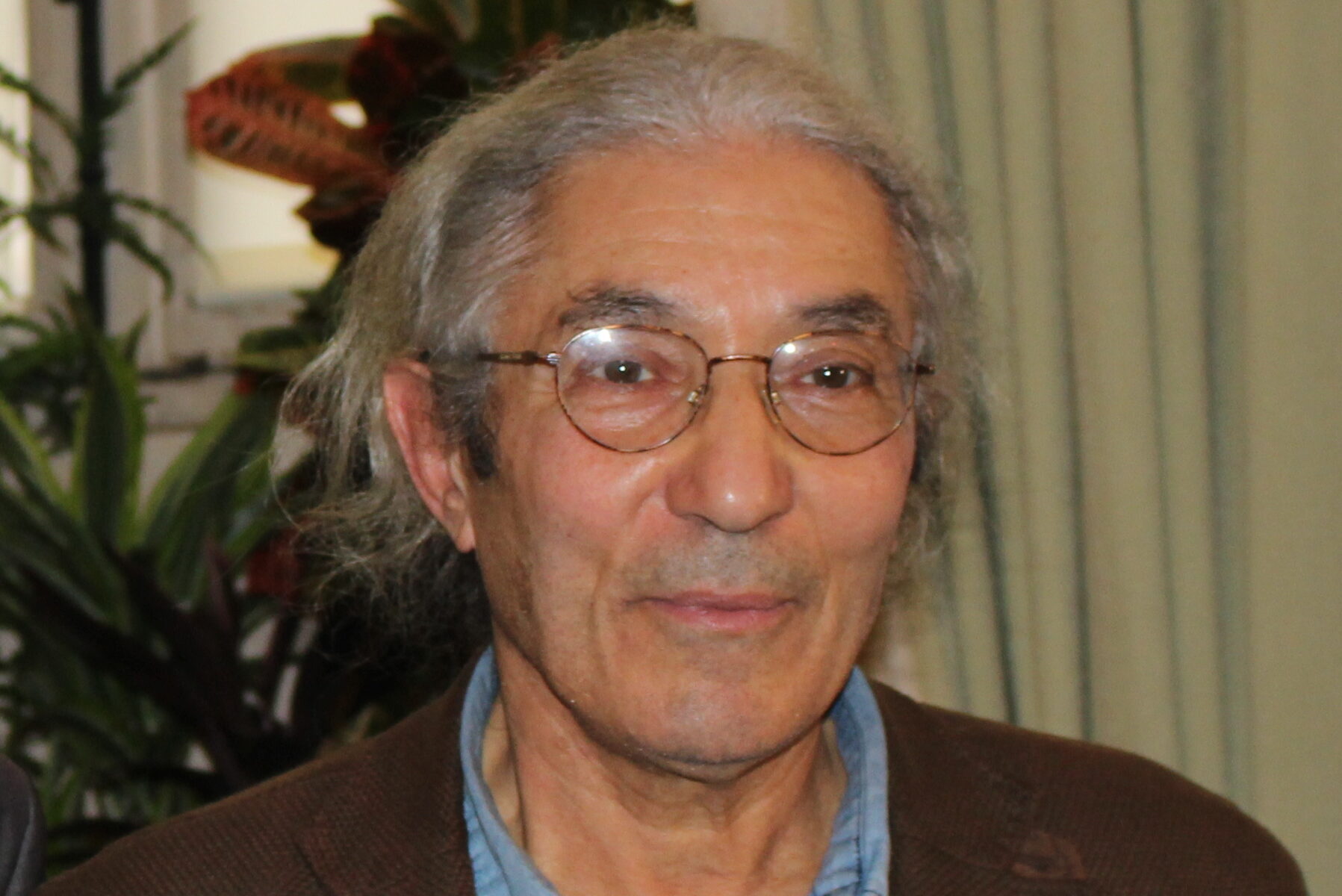 Man with gray hair tied back, wearing glasses, a brown jacket, and a light blue shirt, standing indoors with plants in the background.