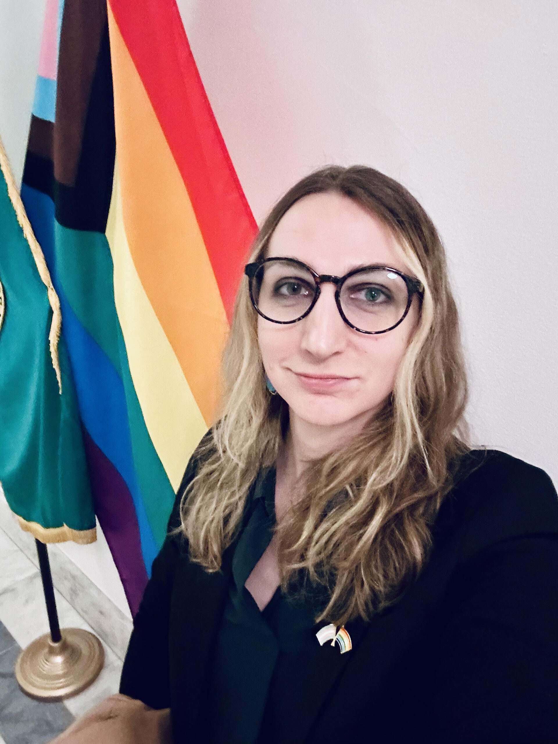 A person with long hair and glasses is taking a selfie indoors. They are wearing a dark blazer with a rainbow pin. A colorful flag with rainbow stripes stands on a pole behind them.