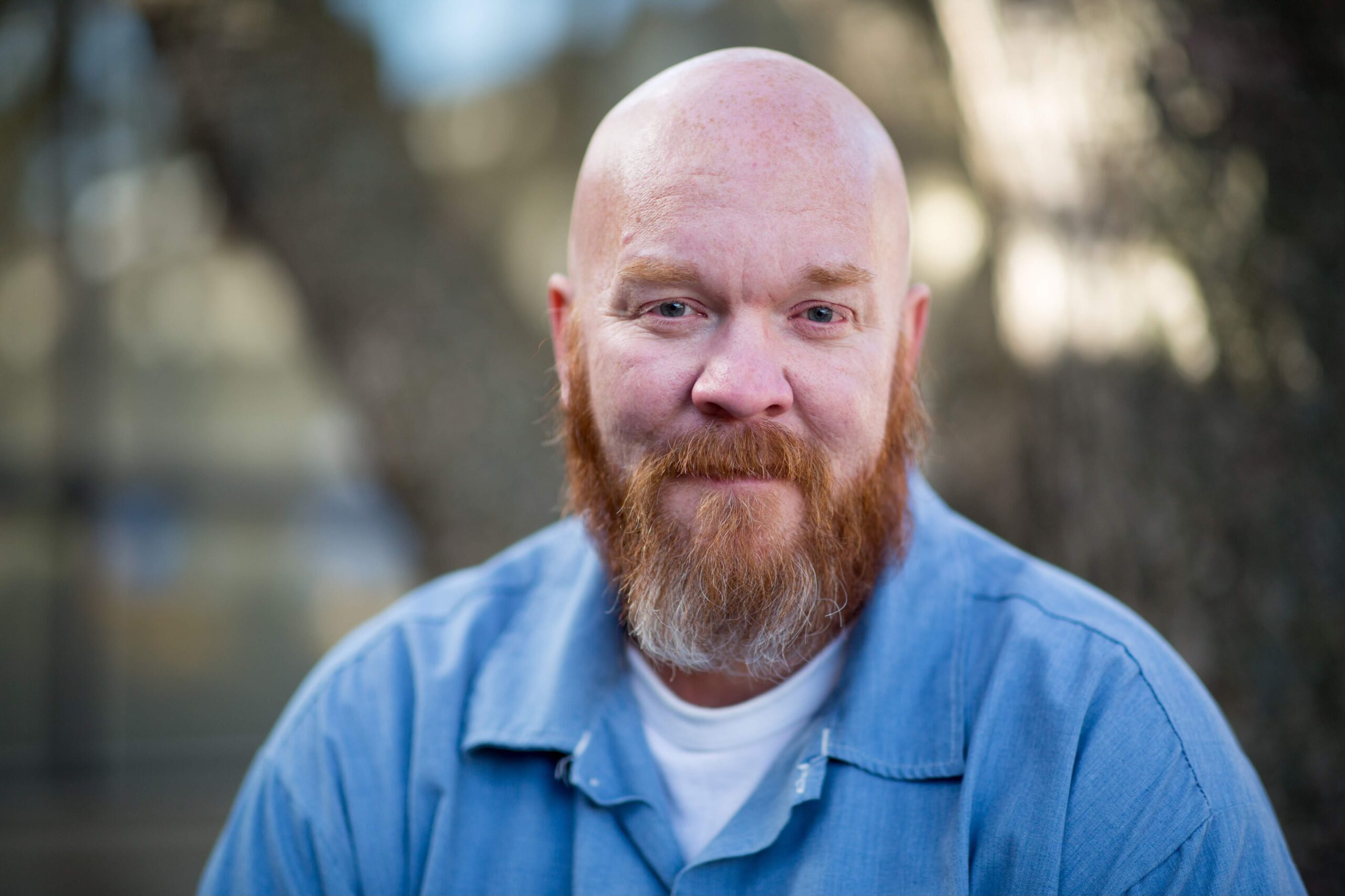 A bearded man with a shaved head, wearing a blue shirt over a white T-shirt, is sitting outdoors with a blurred background of trees.