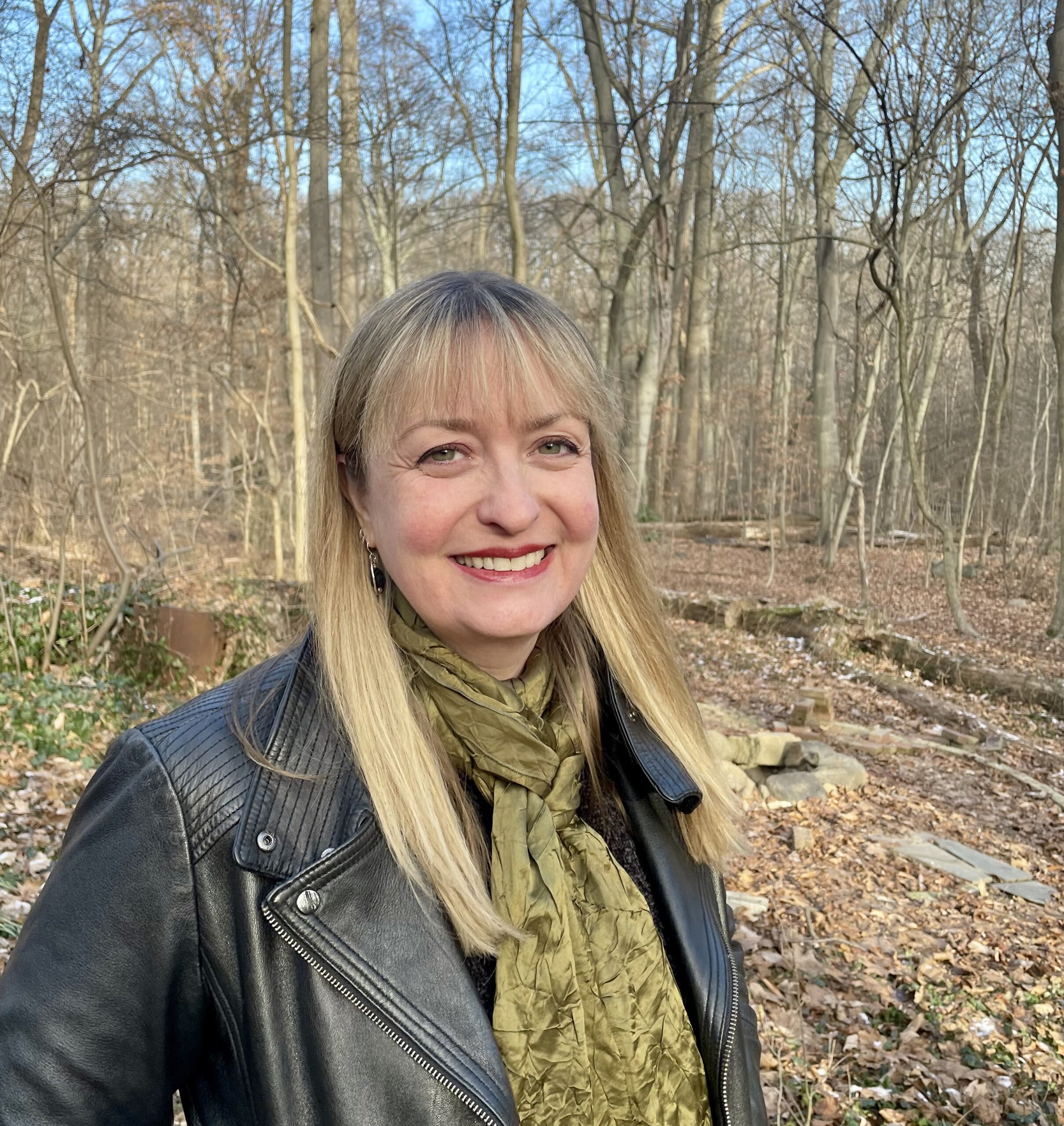 A woman with long blonde hair, wearing a leather jacket and a green scarf, smiles while standing in a forest with bare trees and fallen leaves. The sky is clear and blue.