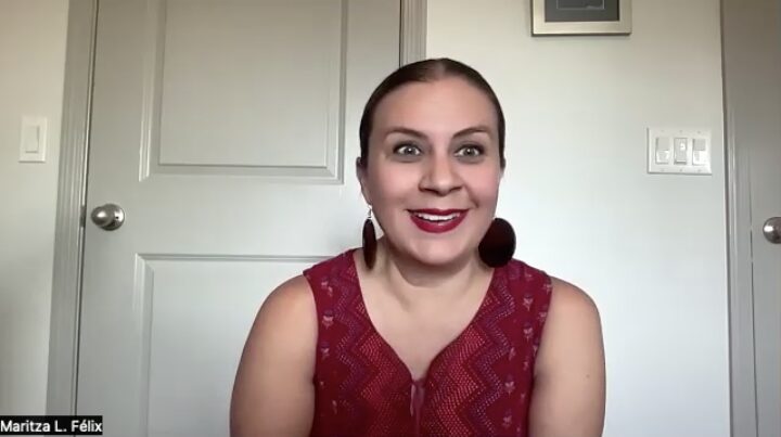 A person with long brown hair is smiling, sitting in front of a closed door. They are wearing a sleeveless red top and large round earrings. A small label on the photo says Maritza L. Félix.