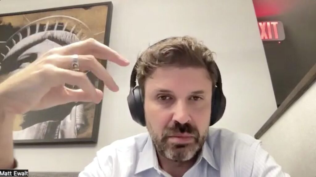 A person wearing headphones is sitting indoors, gesturing with one hand. Behind them is a framed picture of the Statue of Liberty. An exit sign is partially visible in the background.