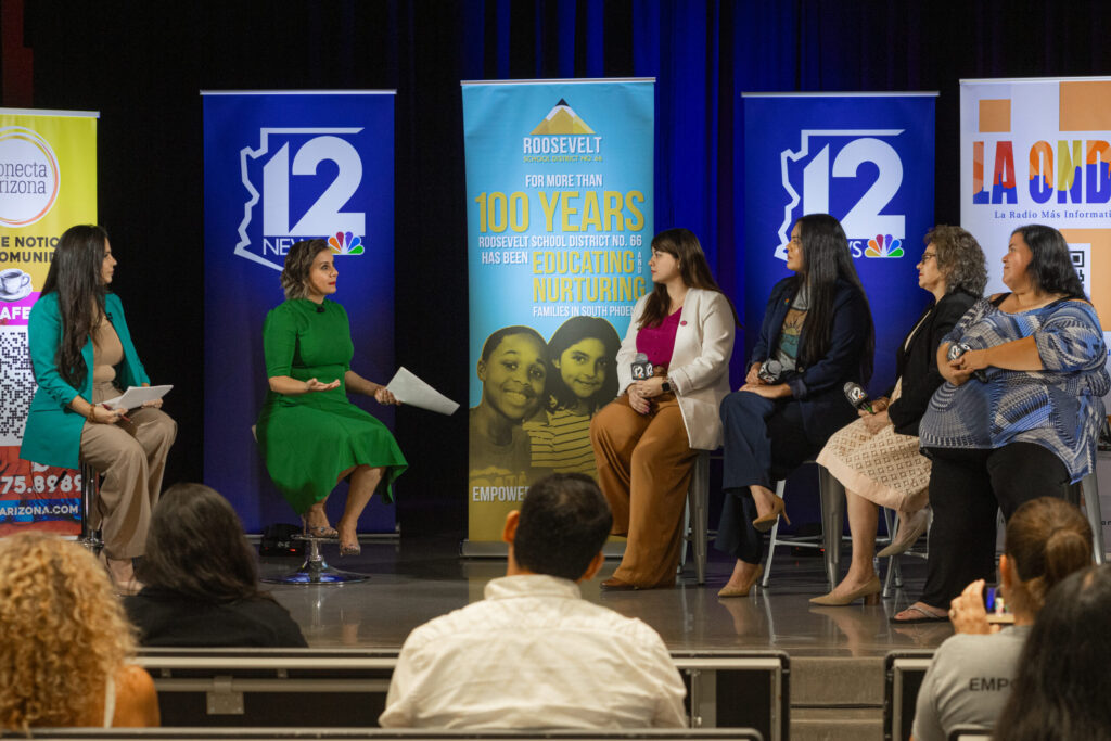A panel of six women sits on stage having a discussion. Behind them are colorful banners, including ones for 12 News and Roosevelt School District. An audience is visible in the foreground.