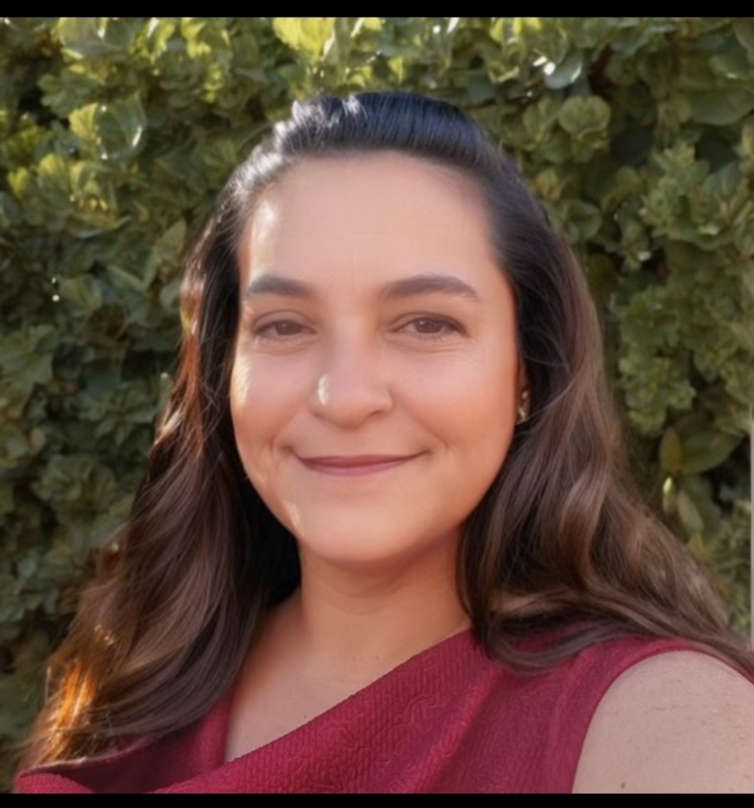 A person with long brown hair and a maroon top smiles at the camera. They are standing outdoors in front of lush green foliage.