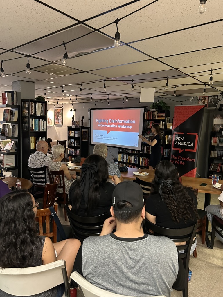 People attending a workshop titled Fighting Disinformation in a room filled with bookshelves. A presenter stands near a screen displaying the workshop title. Participants are seated around tables.