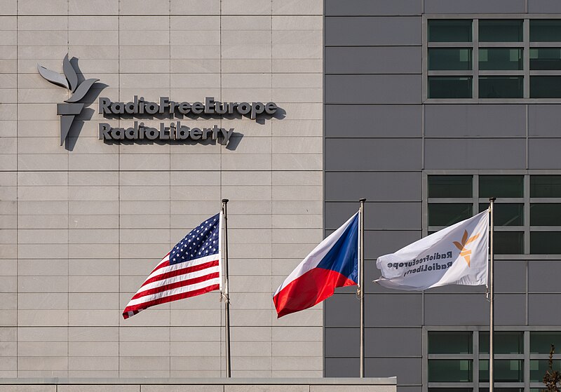 Building with Radio Free Europe Radio Liberty logo. Three flags are in front: American, Czech, and a white flag with the Radio Free Europe logo. The facade is made of rectangular light-colored panels.