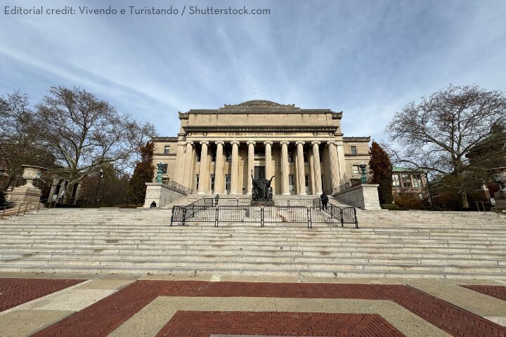 Front view of a neoclassical building with a row of tall columns, set atop a wide staircase. Leafless trees flank the structure, and a cloudy sky forms the backdrop. Brick and stone pathways lead to the steps.