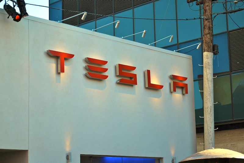 The image shows the exterior of a building with the Tesla logo in large red letters on a white wall. Vertical windows with blue tinted glass are visible, along with a utility pole on the right.