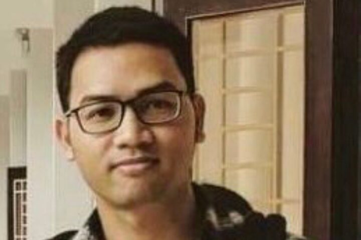 A person with short dark hair and glasses is standing indoors, wearing a dark jacket over a checkered shirt. The background includes a door with glass panels.