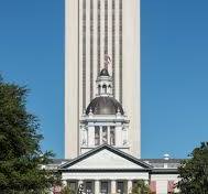 A tall modern building behind a historic white and gray capitol building with a dome. An American flag flies atop the dome. The sky is clear and blue, with trees partially visible on the left side.