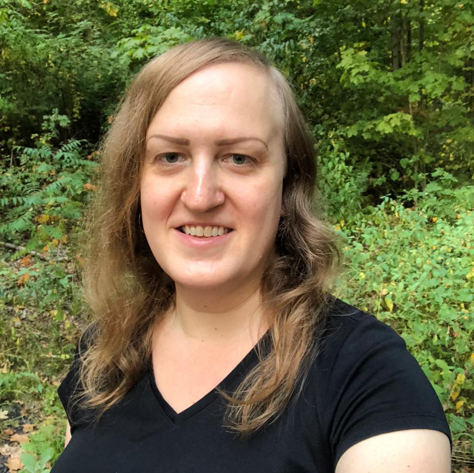 Person with long hair and a black shirt smiling outdoors, standing in front of lush green foliage. The background reveals a blend of leaves and trees.