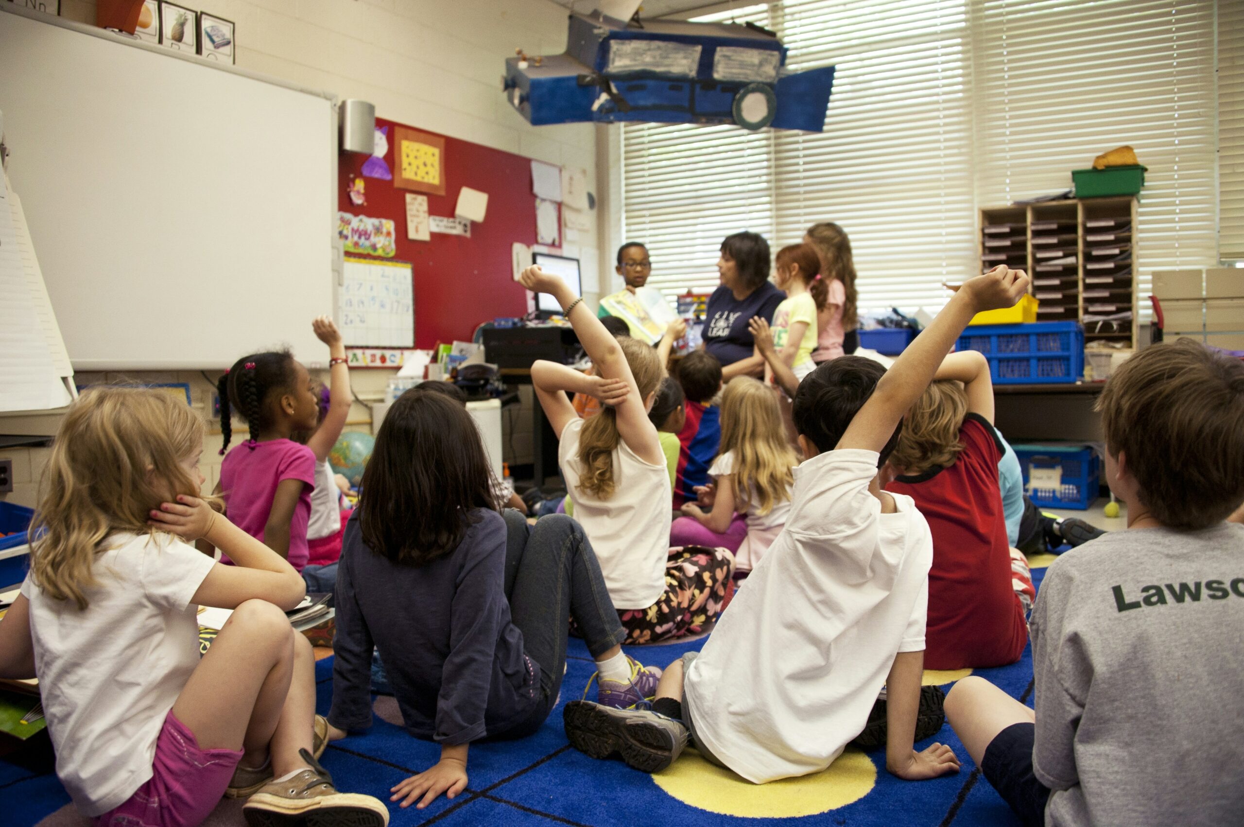 A diverse group of young children sit on a colorful rug in a classroom, some raising their hands. A woman sits at the front, engaging with the students. Bright light filters through the windows, illuminating the scene.
