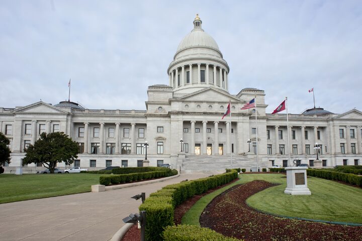 A grand white building with a large central dome sits beneath a cloudy sky. The structure features tall columns and staircases leading to the main entrance. Flags fly atop the building, and there are neatly trimmed lawns and paths in the foreground.