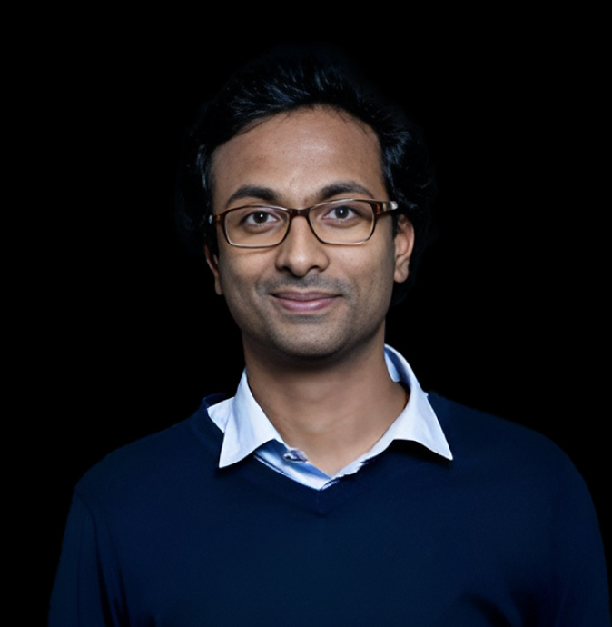 A person with dark hair and glasses, wearing a light blue collared shirt under a dark sweater, smiles while standing against a black background.