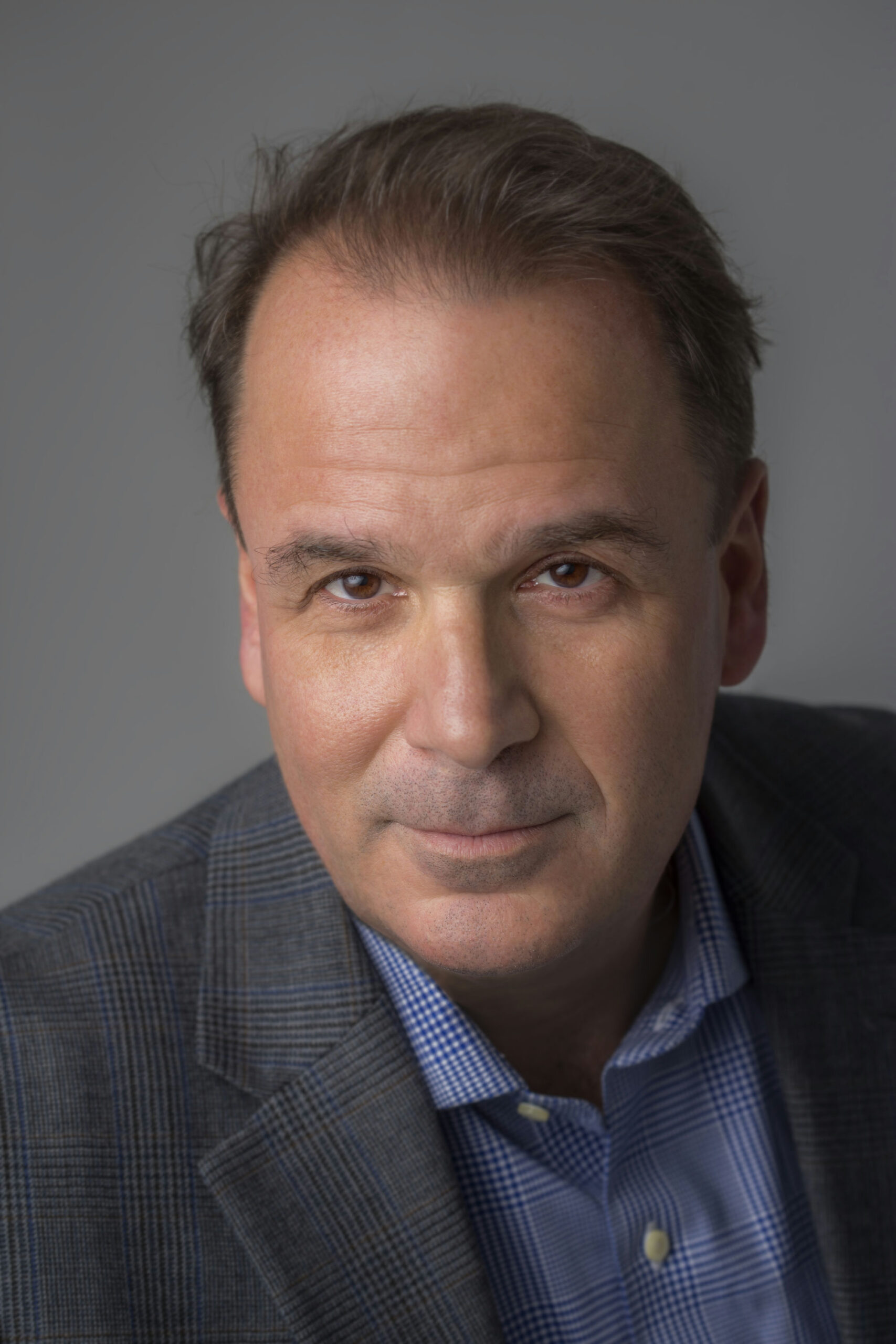 A man with short, dark hair wearing a gray plaid suit jacket and blue checkered shirt, looking directly at the camera against a gray background.