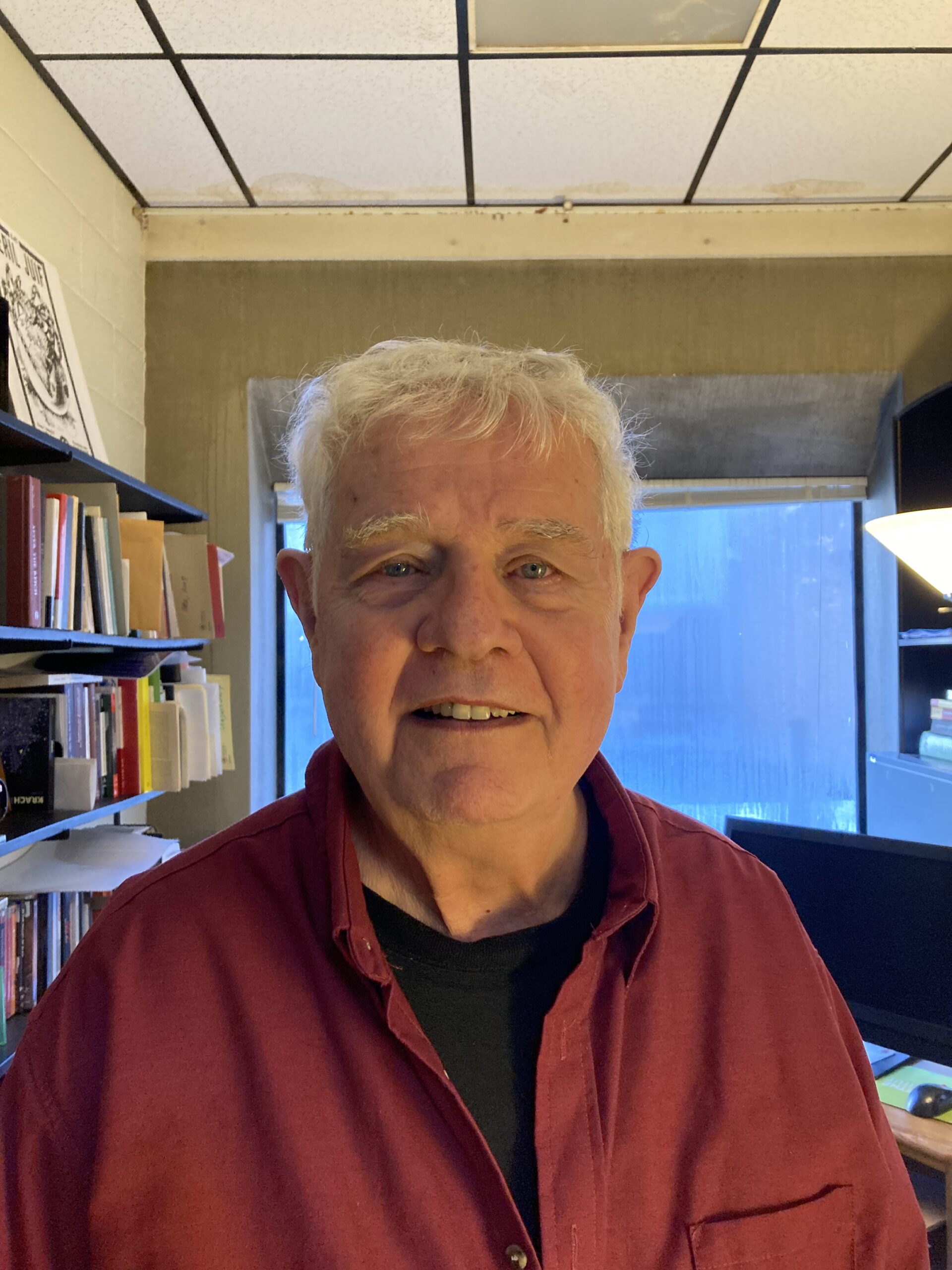 An older man with white hair, wearing a red shirt over a black t-shirt, stands in an office filled with bookshelves and a computer. The room has a window with a partially blurred outdoor view.