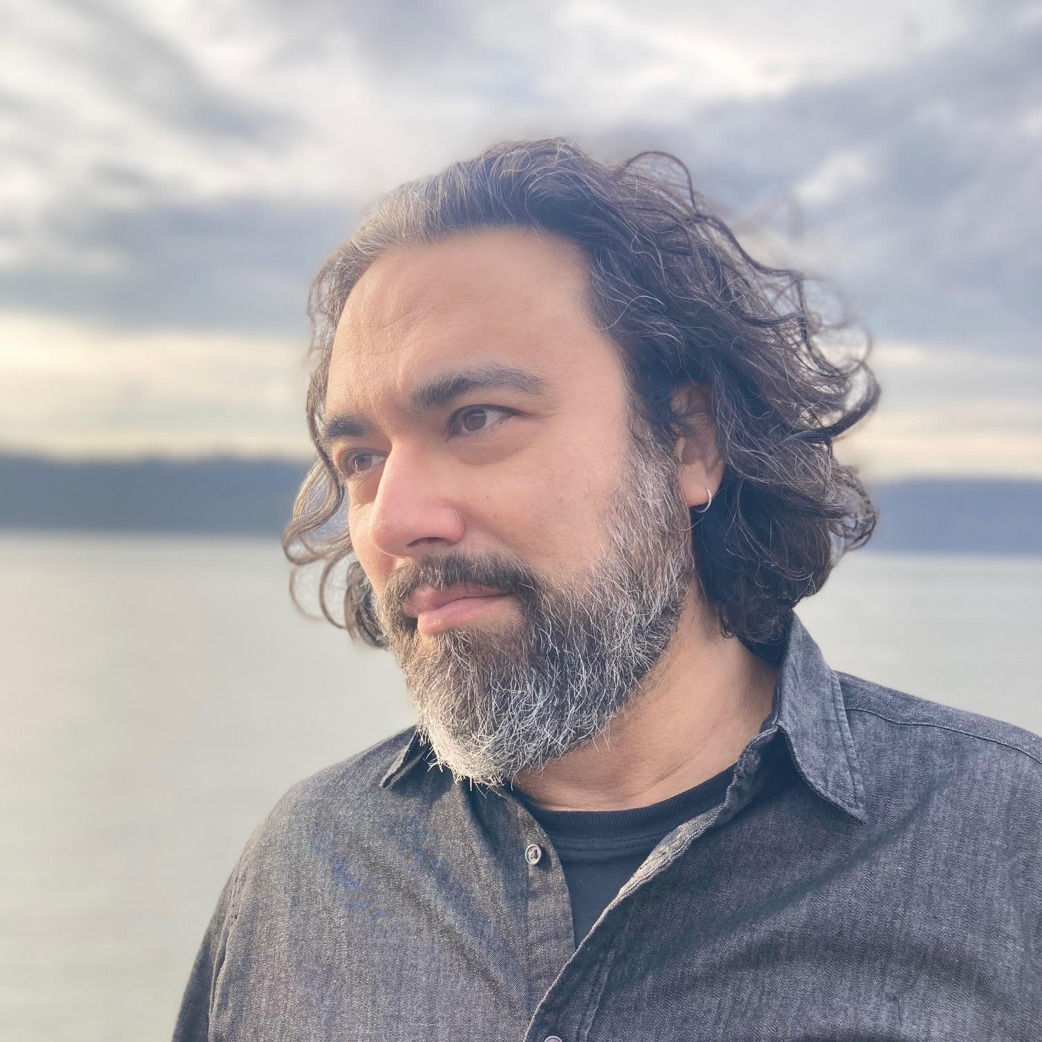 A man with shoulder-length wavy hair and a beard stands in front of a calm body of water, wearing a dark button-up shirt. The background features a cloudy sky and distant hills.