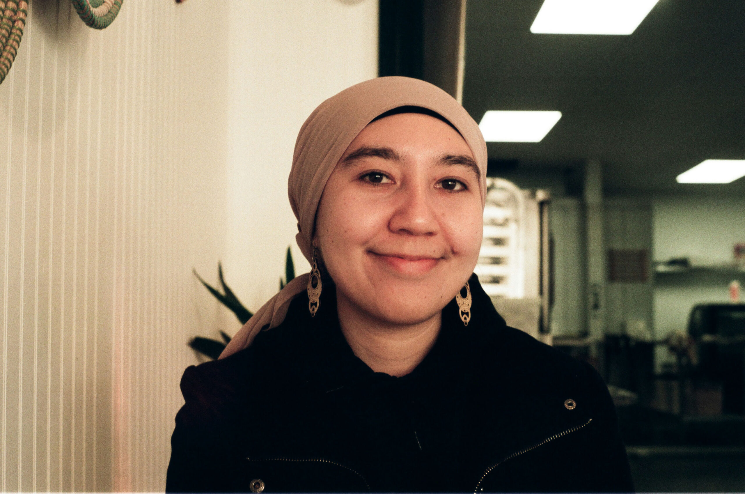A person wearing a beige headscarf and dark clothing smiles at the camera in an indoor setting. The background includes white walls, a few plants, and fluorescent lighting.