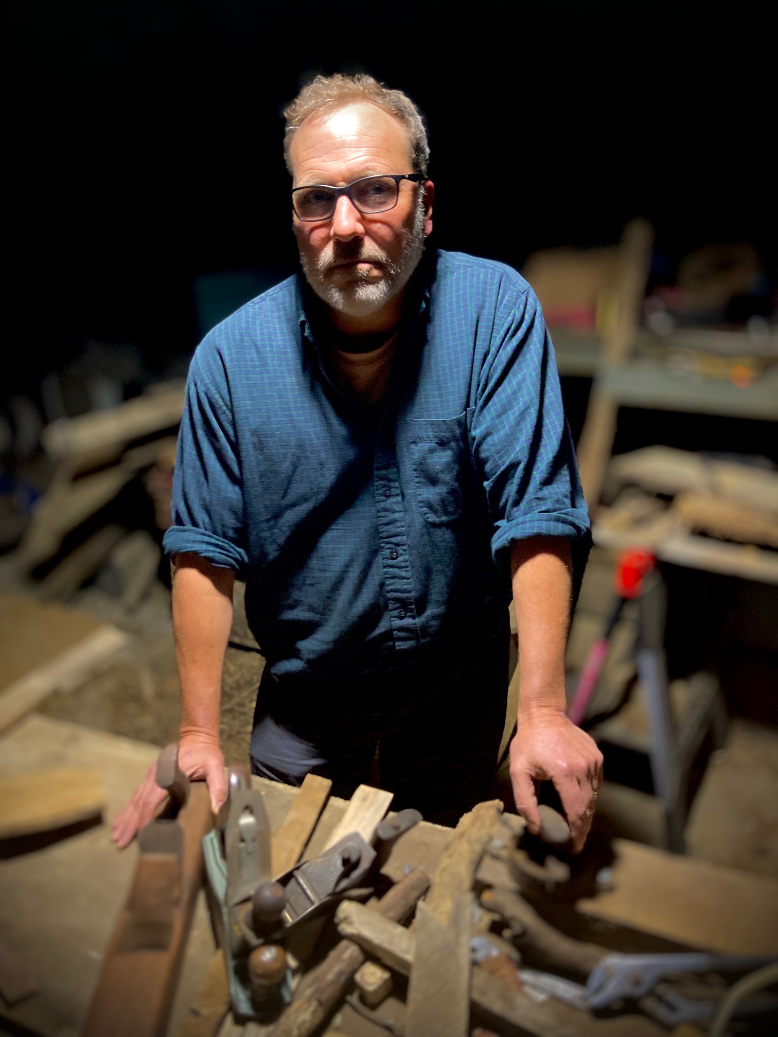 A man with glasses and a beard stands at a workbench, wearing a blue shirt. He is surrounded by various carpentry tools in a dimly lit workshop. The focus is on his serious expression and the tools in front of him.