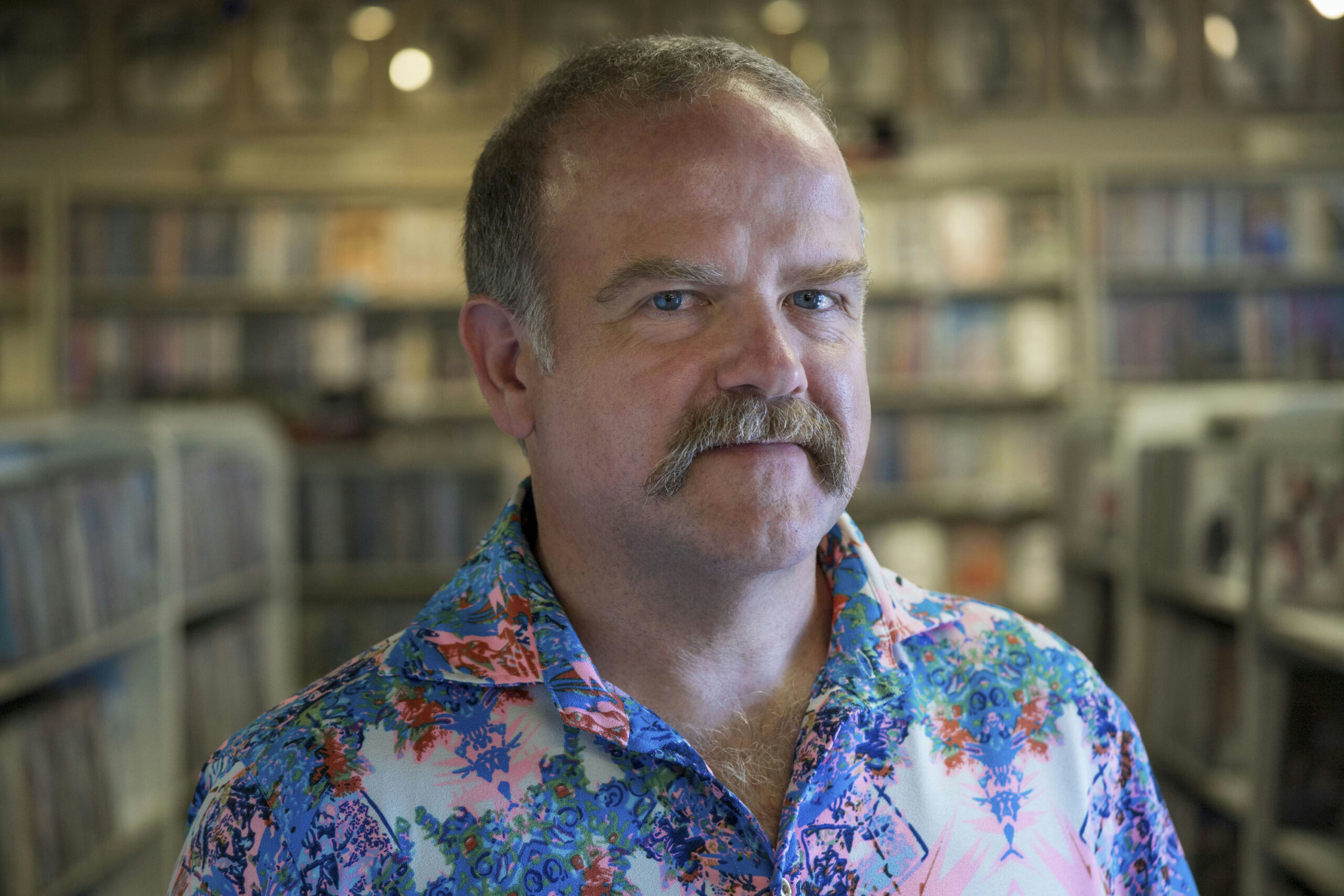 A man with a mustache wearing a colorful floral shirt stands in a store filled with shelves of vinyl records. The background is slightly out of focus, highlighting the records and creating a nostalgic atmosphere.