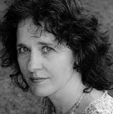 A black and white portrait of a person with curly hair, looking towards the camera. They are wearing a necklace and earrings, with a soft, natural outdoor background.