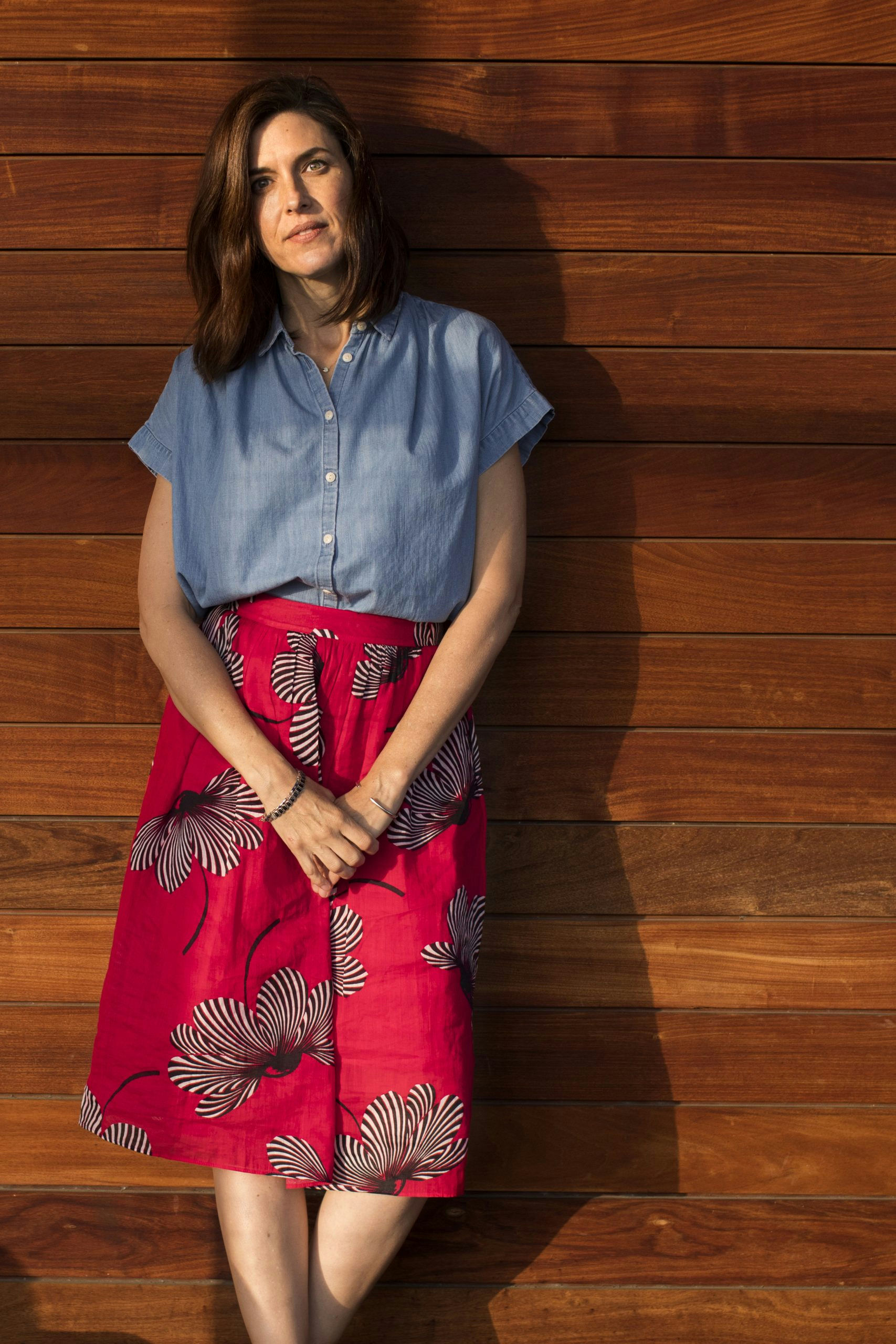 A woman stands against a wooden wall, looking into the distance. She wears a blue short-sleeved shirt and a red skirt with a white floral pattern. Her hair is shoulder-length and brown, and soft sunlight creates a natural shadow behind her.