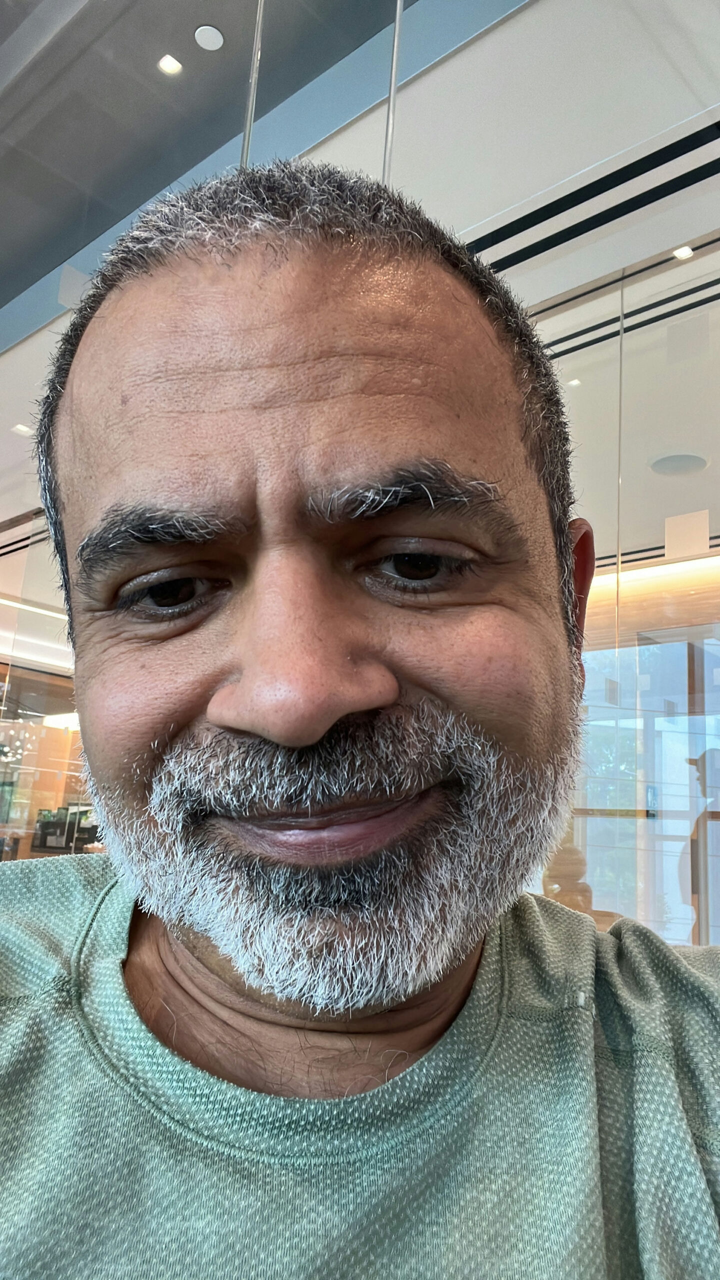 A person with short, graying hair and a beard is smiling at the camera. They are indoors, wearing a green shirt, with glass walls and modern interior elements visible in the background.