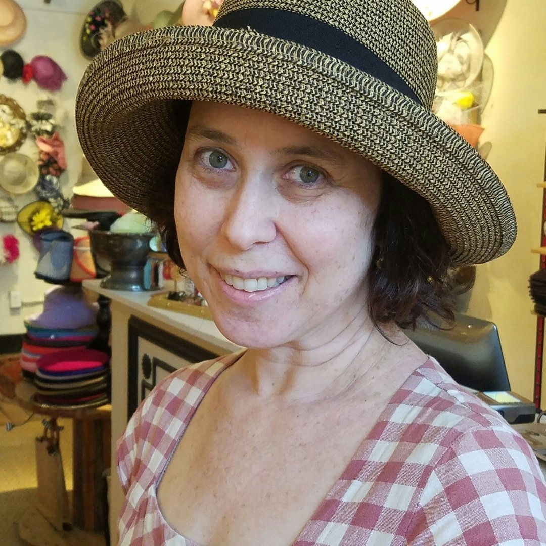 A woman in a plaid dress smiles while wearing a wide-brimmed straw hat with a black band. Background shows various colorful hats on display in a shop.