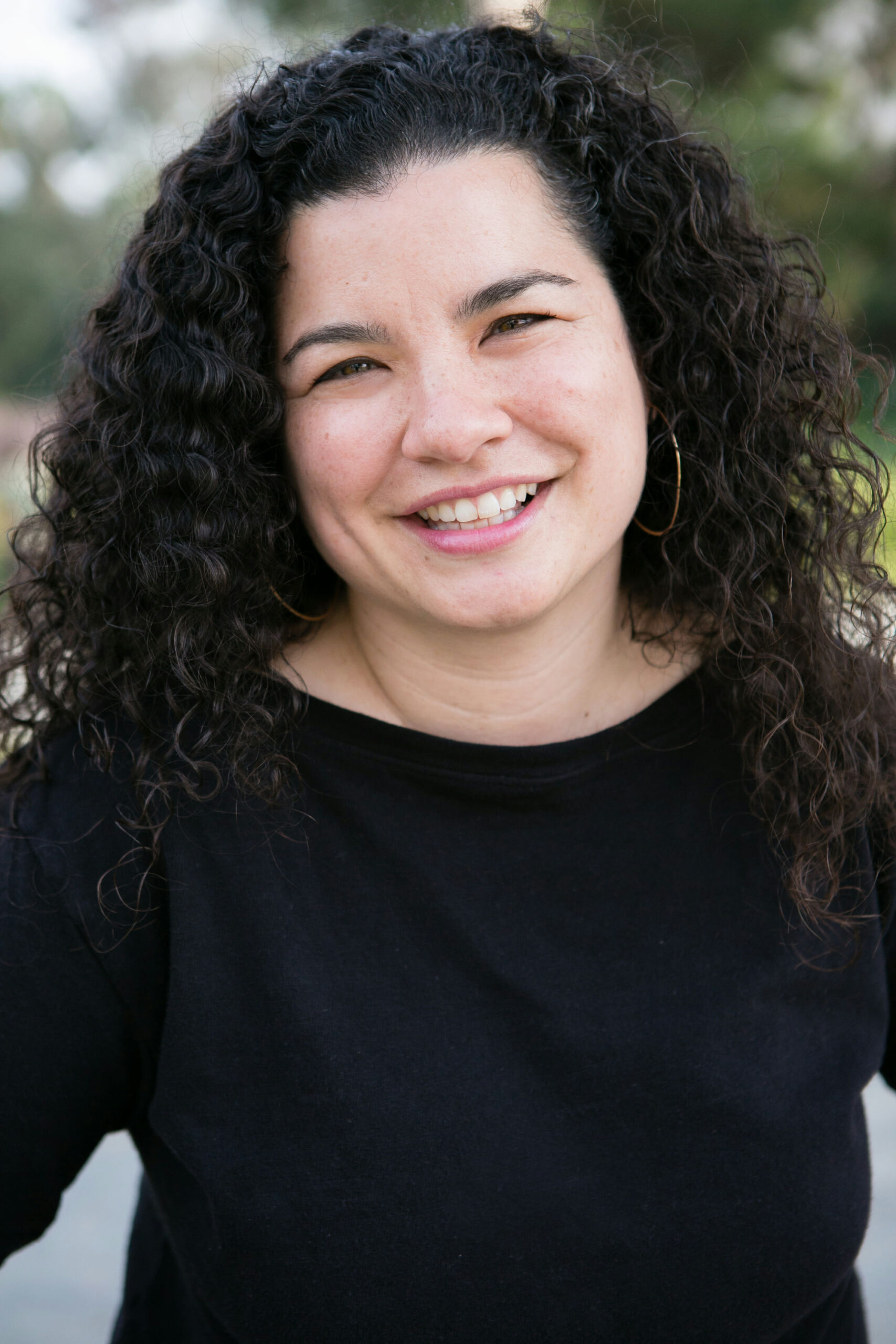 A person with long, curly dark hair is smiling at the camera. They are wearing a black top and are outdoors, with blurred greenery in the background.