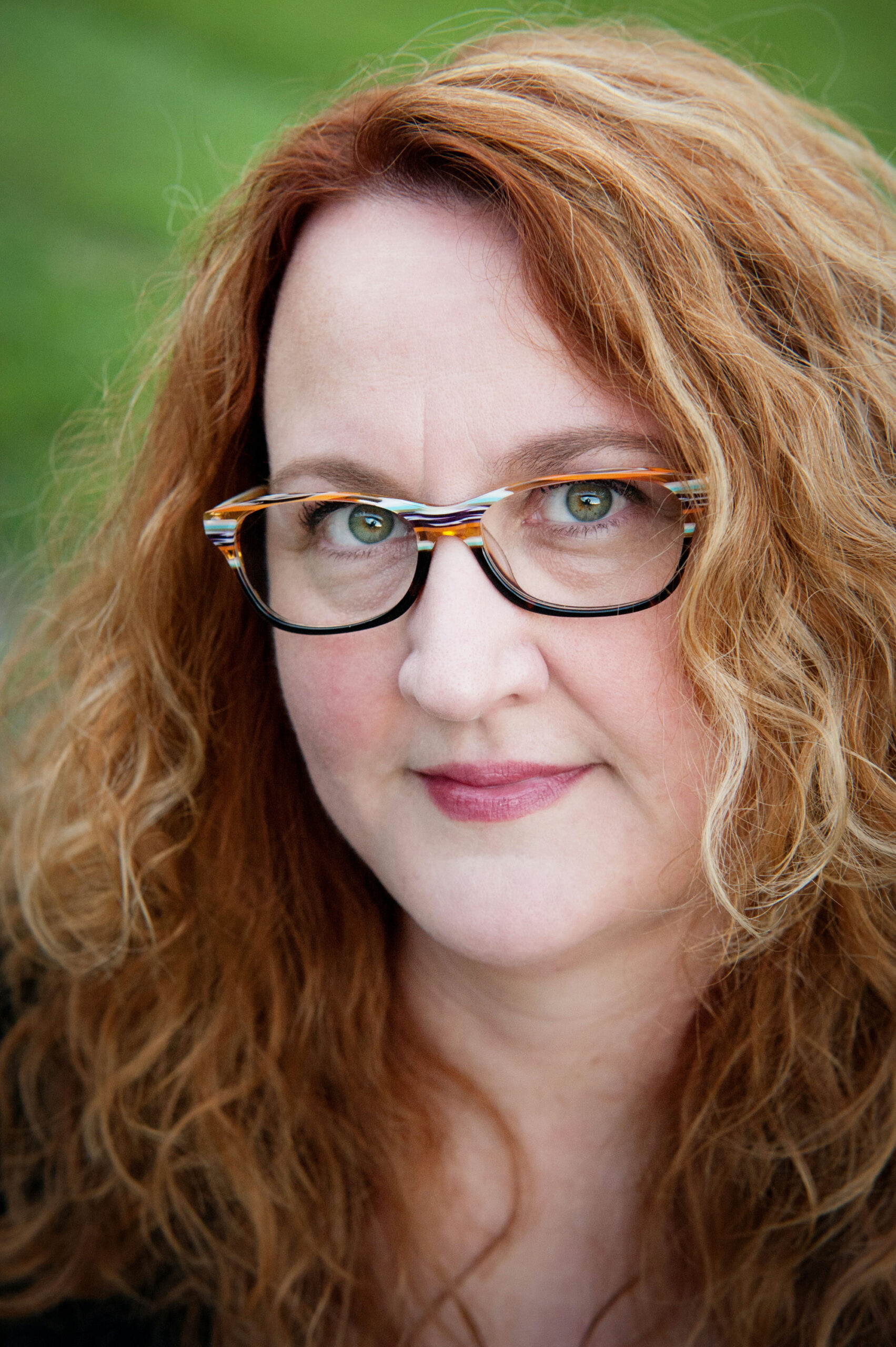 A woman with long, curly red hair and glasses looks directly at the camera. She is outdoors, with a blurred green background, and wears a subtle expression.