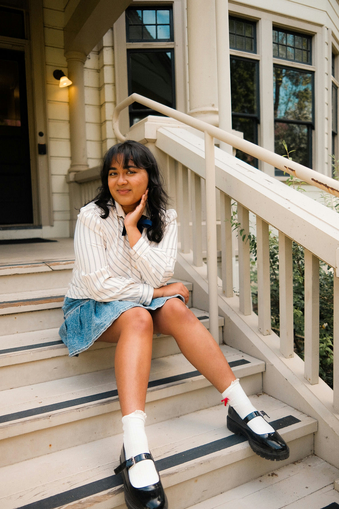 A person with dark hair sits on porch steps, wearing a striped shirt, denim skirt, white socks, and black shoes. They are looking at the camera with a relaxed expression. The house in the background is light-colored with dark window frames.