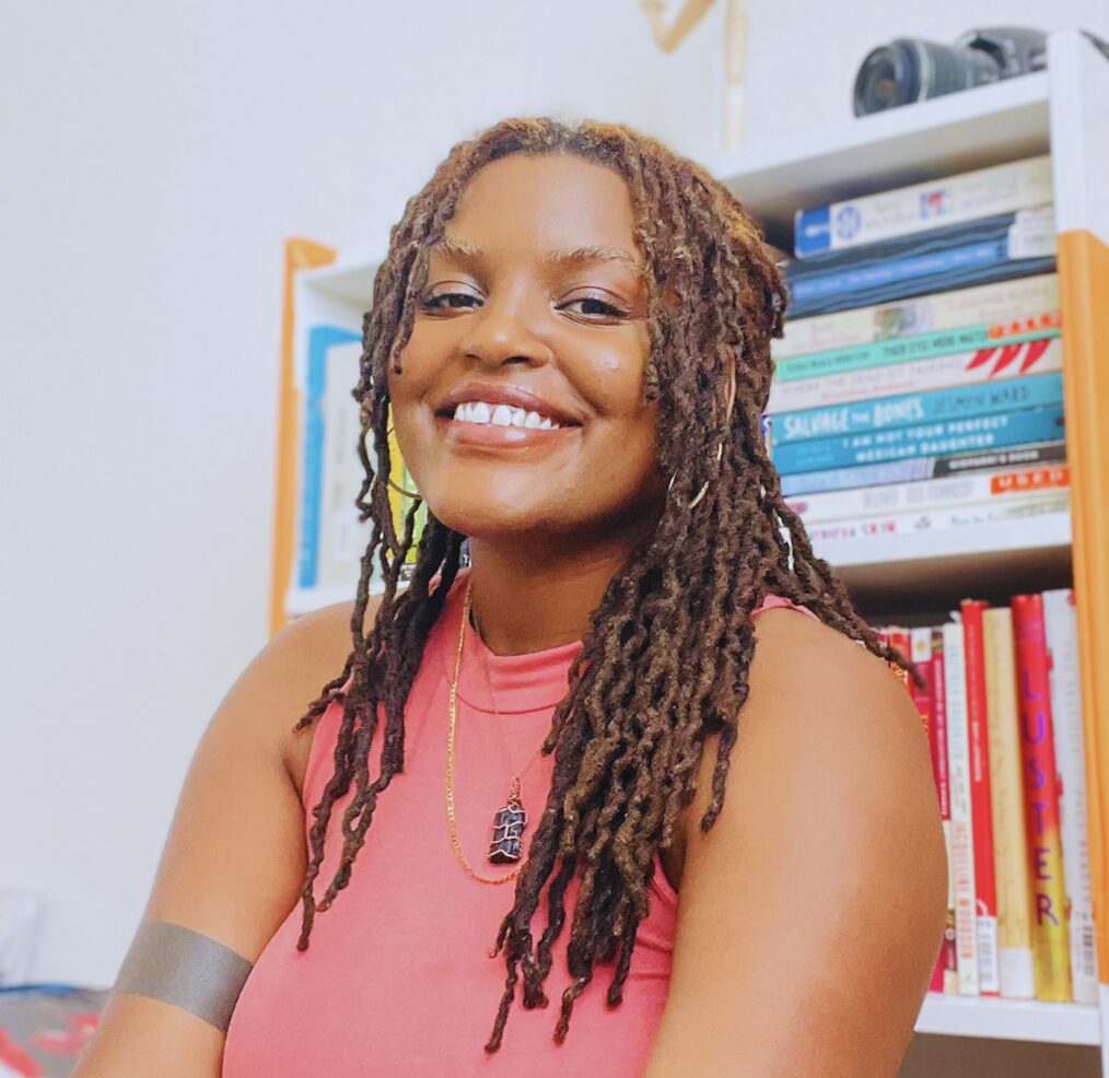Smiling person with long braids sits in front of a bookshelf filled with various books. They wear a pink sleeveless top, a necklace, and a dark armband. The background shows a camera on the top shelf.