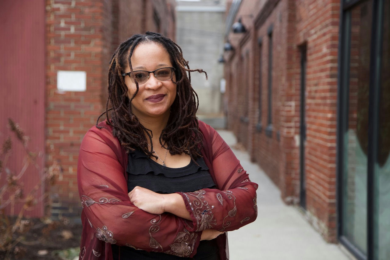A person with glasses and long hair stands smiling with arms crossed in an alleyway between brick buildings. They wear a sheer red shawl over a black top. The buildings have windows and there is a blurry background.