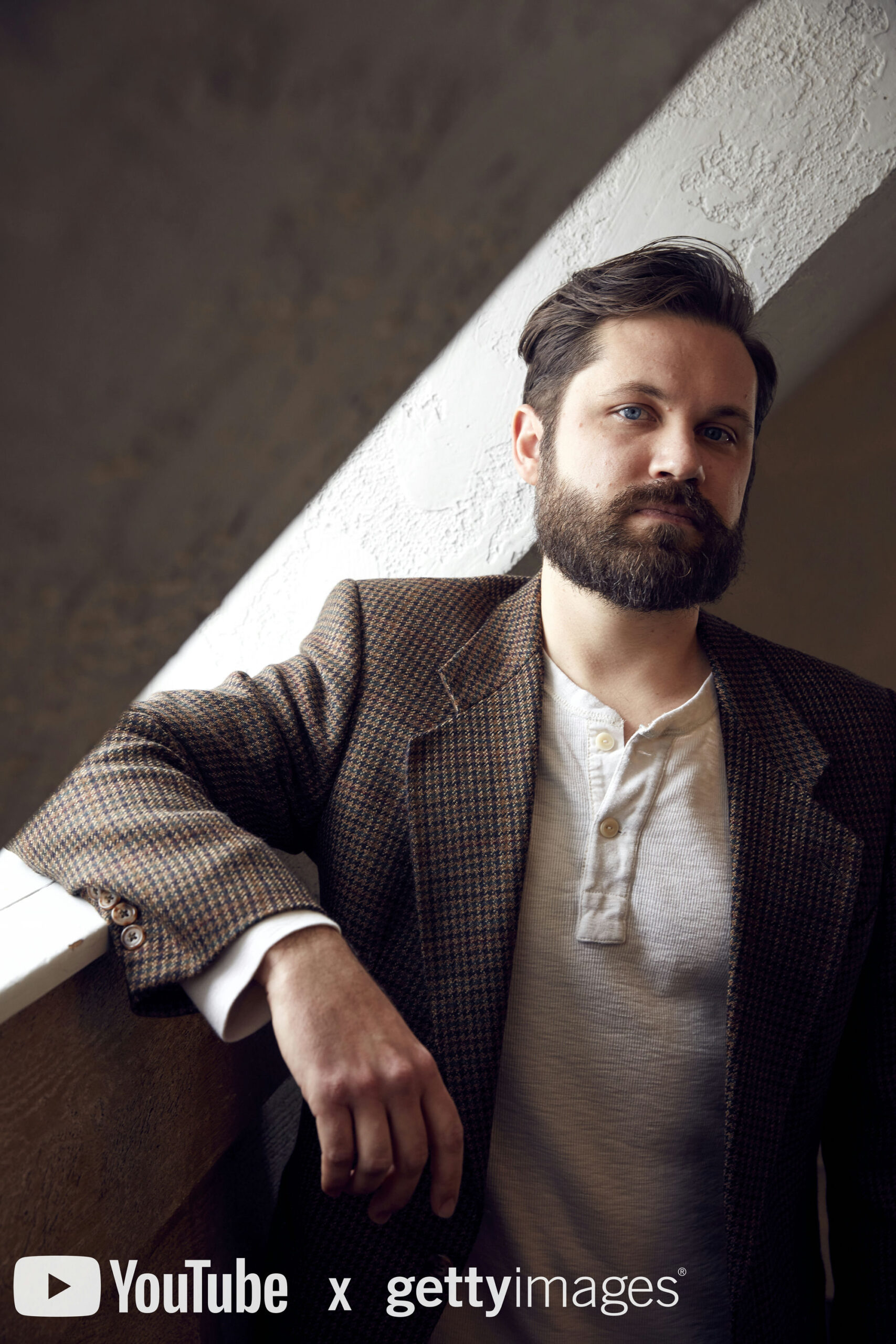 Bearded man with dark hair leaning against a white wall, wearing a checkered blazer over a light henley shirt. Sunlight casts a diagonal shadow. Visible textures on wall and clothing. YouTube and Getty Images logos in the bottom corner.