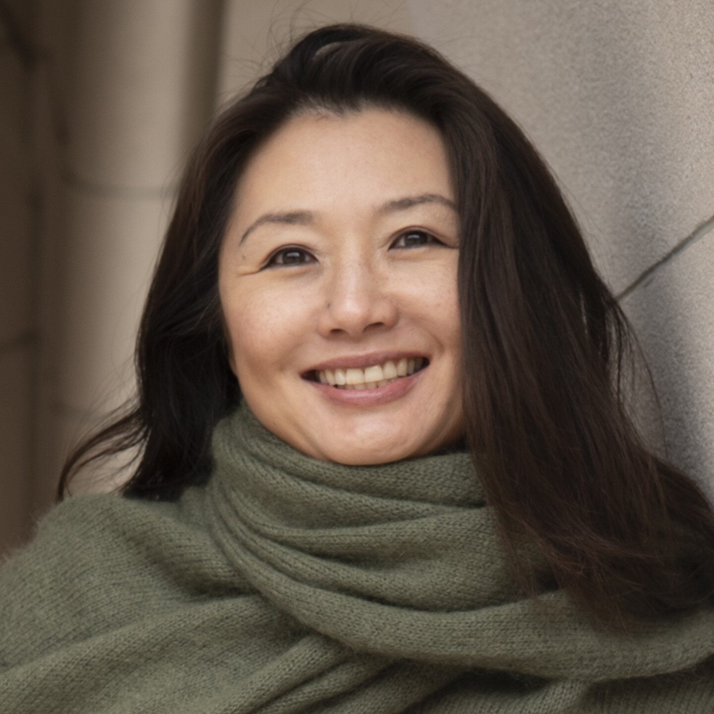 A woman with long dark hair and an olive green scarf smiles warmly while leaning against a stone wall.