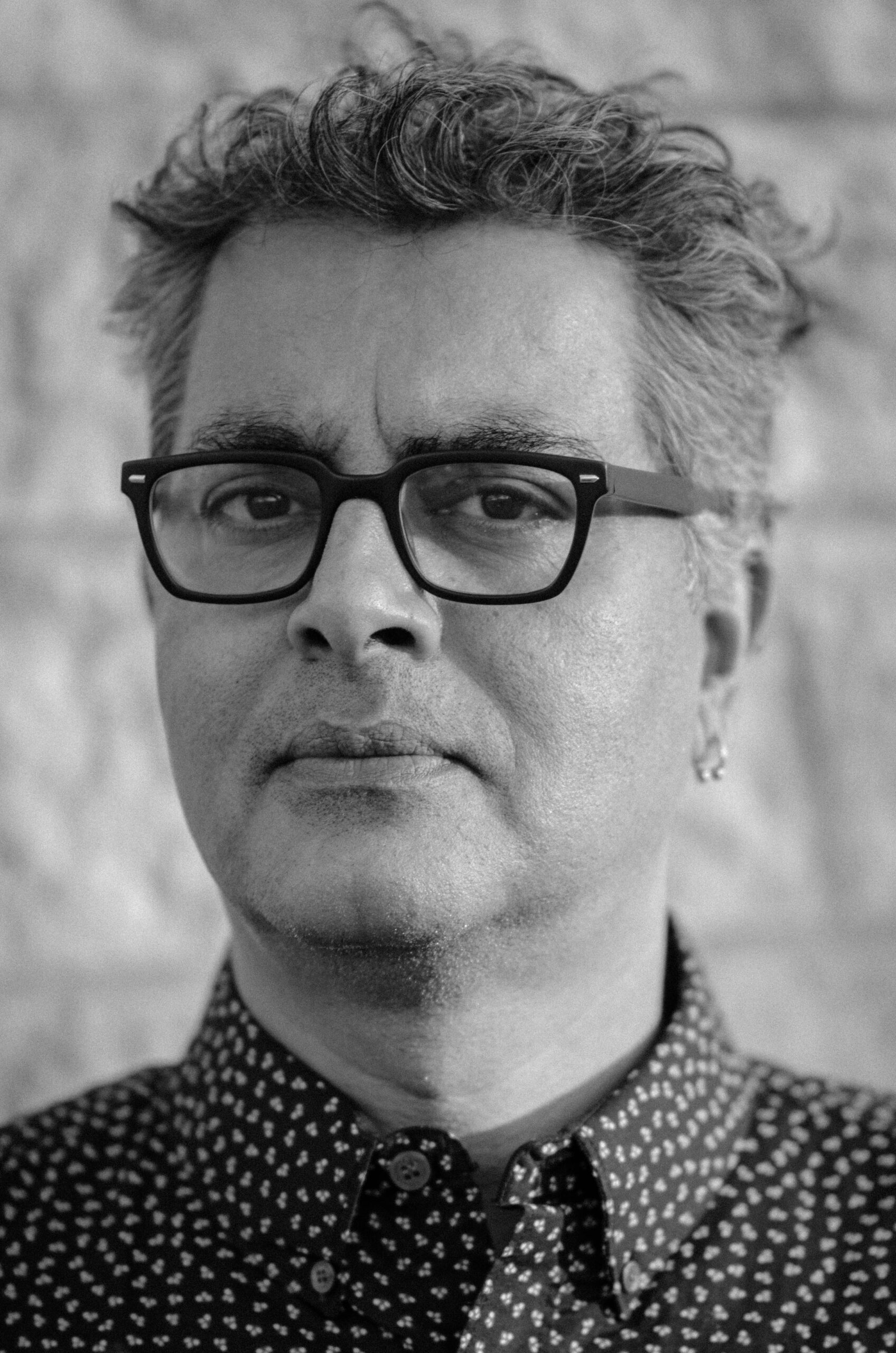 Black and white portrait of a person with short curly hair, wearing dark-rimmed glasses, a patterned button-up shirt, and a small earring, standing in front of a light-colored stone wall.