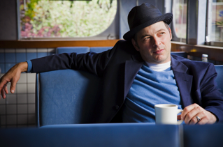 A man wearing a hat and a blazer sits in a blue booth at a diner, holding a white coffee mug and gazing thoughtfully out the window.