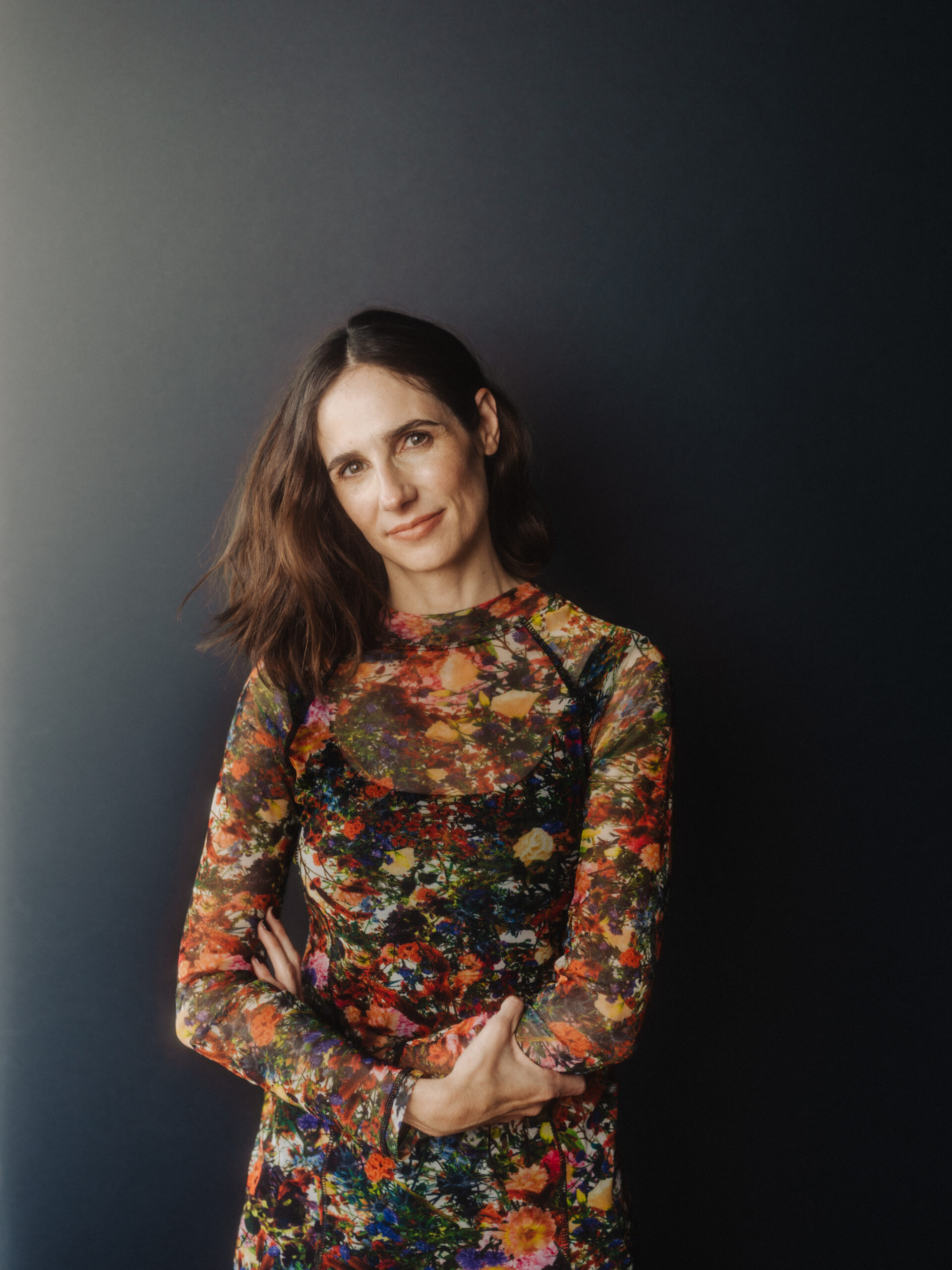 A woman with long brown hair stands against a dark background, wearing a colorful floral-patterned dress with sheer sleeves. She has a relaxed expression and her arms are crossed gently.
