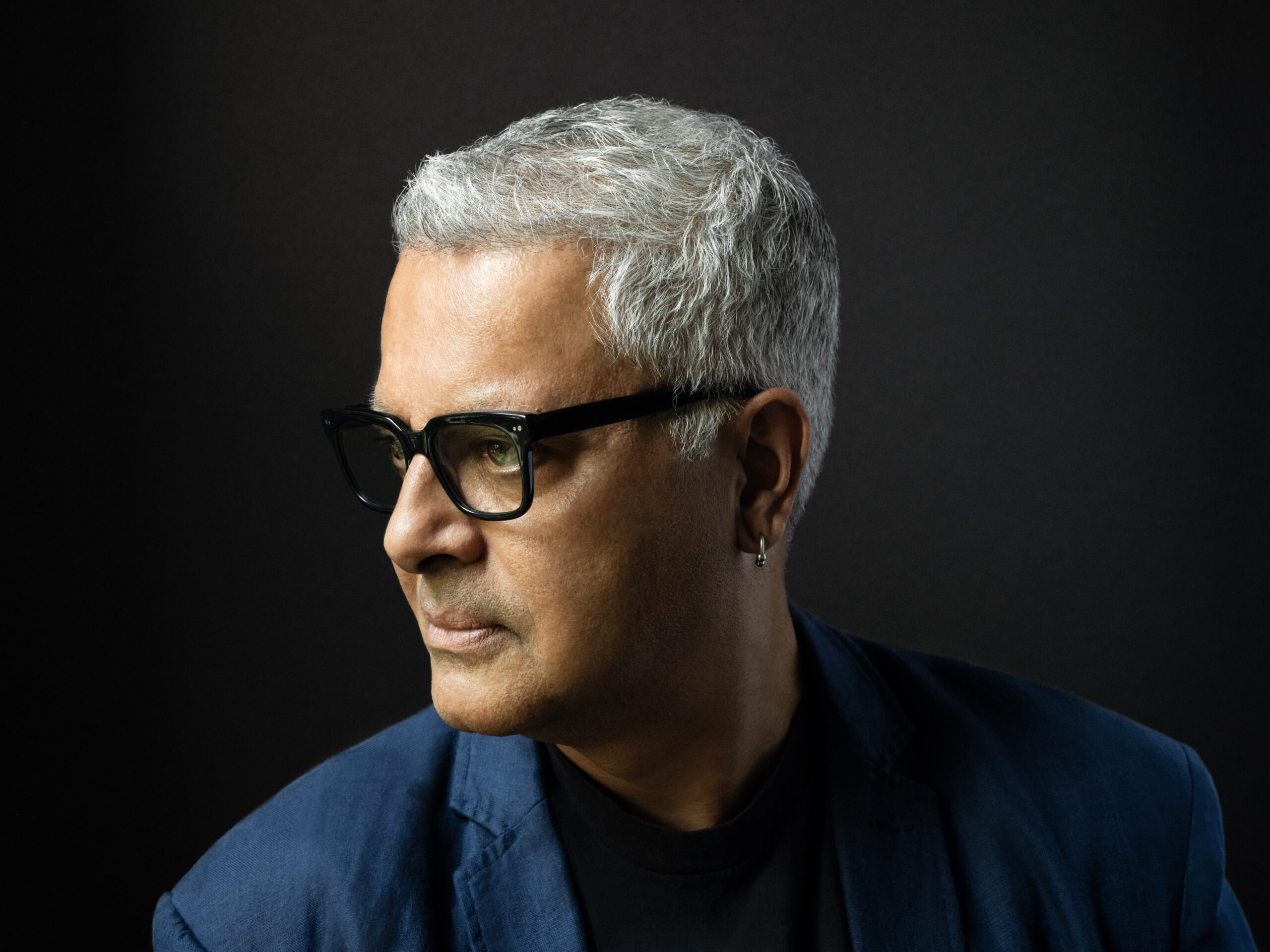 A man with short gray hair, wearing black glasses, a dark shirt, and a blue blazer, looks thoughtfully to the side against a dark background.