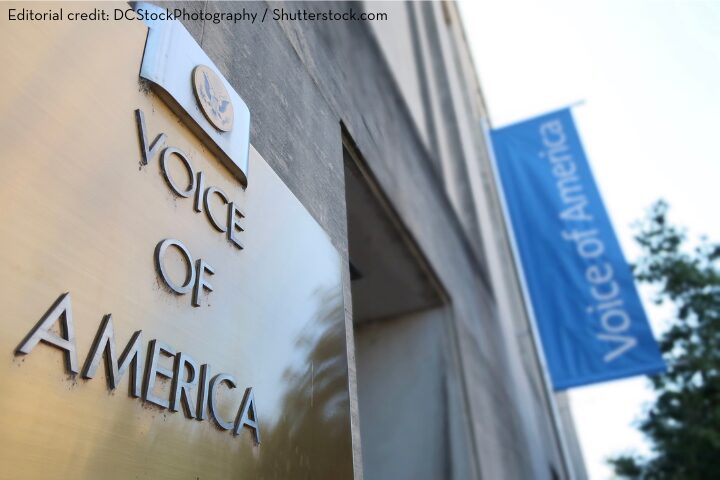 A metal sign on a building reads Voice of America. A blue vertical banner with the same text hangs nearby. The image is credited to DCStockPhotography/Shutterstock.com.