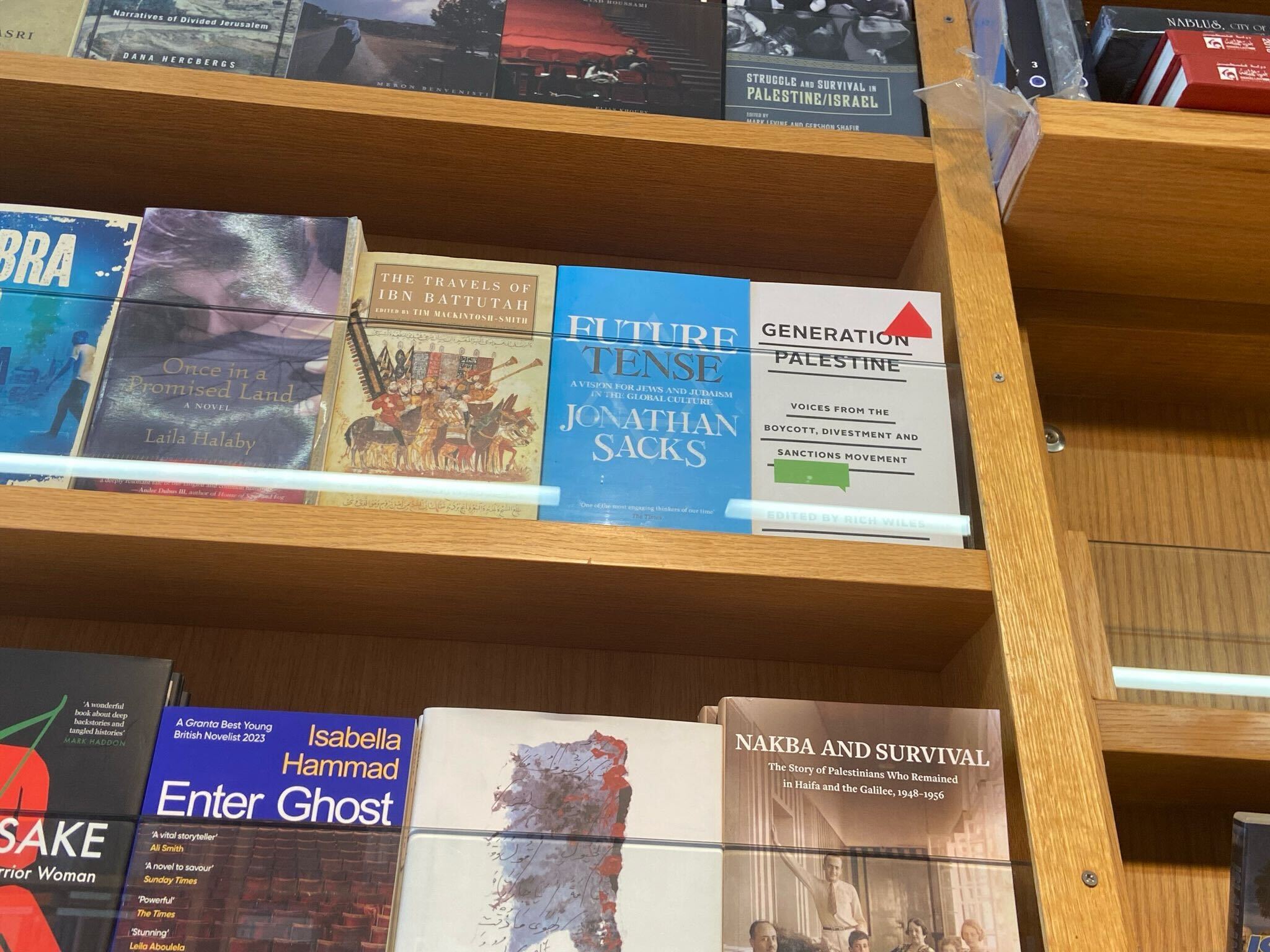 Books displayed on wooden shelves in a bookstore, including titles such as Future Tense by Jonathan Sacks, Generation Palestine, The Travels of Ibn Battutah, and Nakba and Survival.