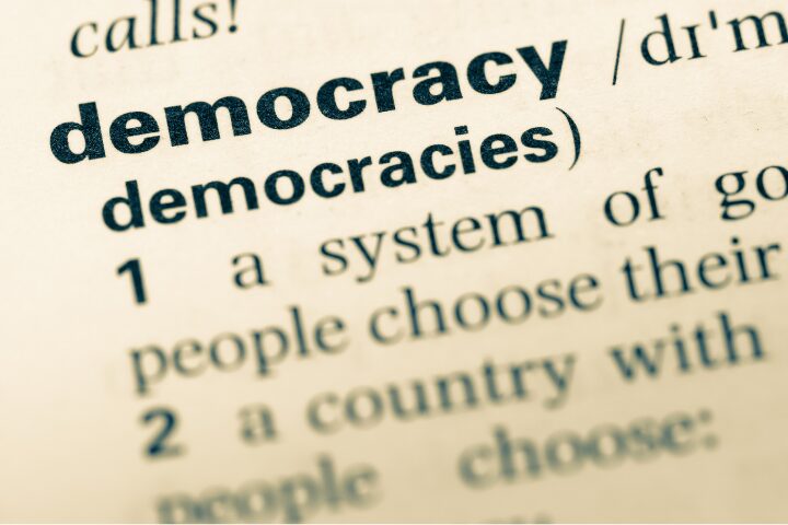 Close-up of a dictionary page showing the word democracy in bold, with its pronunciation and part of the definition visible underneath.