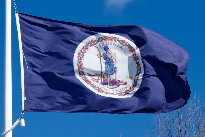 The Virginia state flag waving against a blue sky, featuring the state seal in the center with a woman standing over a defeated figure and the words Sic Semper Tyrannis below.