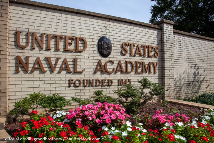 A brick wall with bronze letters reading United States Naval Academy Founded 1845 surrounded by red, pink, and white flowers and green plants.